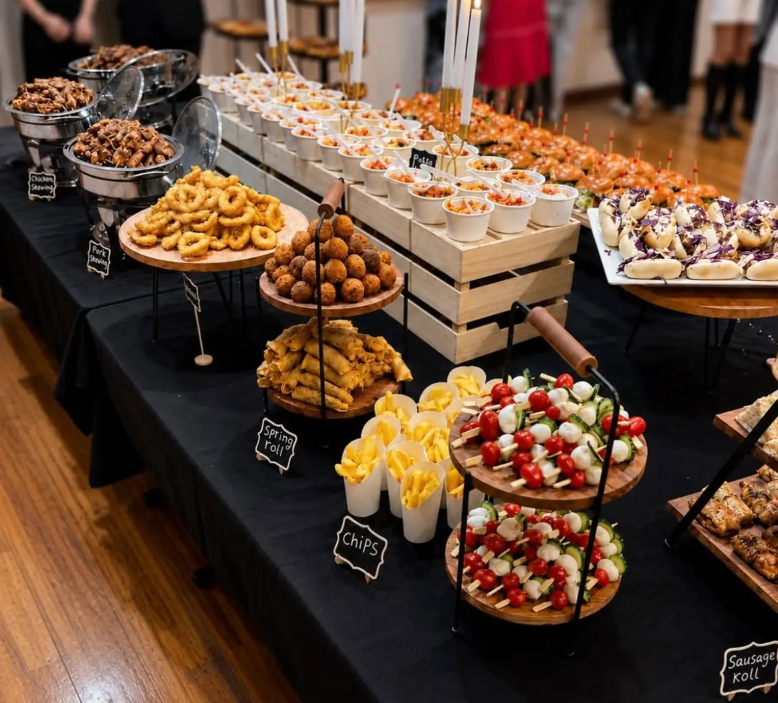 Long buffet table with chicken and pork skewers, calamari, arancini, spring rolls, chips and caprese skewers